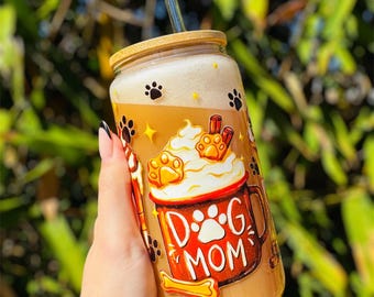 May include: A glass tumbler with a bamboo lid and a clear straw, filled with a coffee-like beverage. The tumbler is decorated with paw prints, stars, and a "Dog Mom" design. The drink has a foamy top.