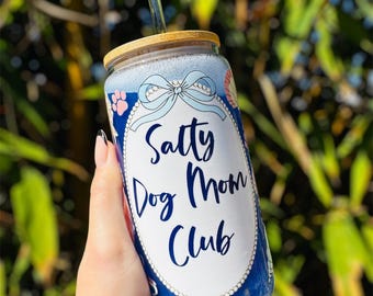 May include: A glass tumbler with a bamboo lid and a clear straw. The tumbler features a blue and white design with the text "Salty Dog Mom Club" in cursive. The design includes a bow and paw prints.