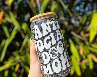 May include: A glitter-covered tumbler with a bamboo lid and a clear straw. The tumbler features the text "ANTI SOCIAL DOG MOM" in white, retro-style lettering, with a paw print graphic. The background is blurred green foliage.