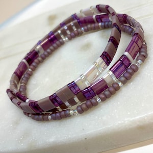 May include: A multi-strand bracelet featuring a mix of purple, mauve, and iridescent beads. The bracelet is composed of rectangular and round beads, creating a layered effect. The design is elegant and suitable for everyday wear.