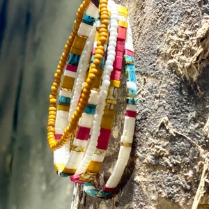 May include: A set of five colorful beaded bracelets in shades of white, yellow, red, and blue. The bracelets are stacked on top of each other and are hanging from a wooden surface.