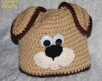 May include: A handmade crochet dog hat in tan with brown trim. The hat features large googly eyes, a black nose, white cheeks, and a pink tongue. The hat is designed to resemble a dog's face.