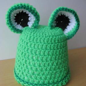 K&ouml;nnte beinhalten: Gr&uuml;ne H&auml;kelm&uuml;tze mit zwei gro&szlig;en Ohren, die an einen Frosch erinnern. Die Ohren sind wei&szlig; und schwarz mit gr&uuml;nem Rand.