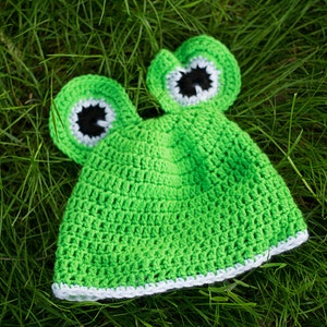 K&ouml;nnte beinhalten: Gr&uuml;ne geh&auml;kelte Frosch-M&uuml;tze mit wei&szlig;em Rand und schwarzen Augen. Die M&uuml;tze liegt auf einem Bett aus gr&uuml;nem Gras.