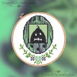 Puede incluir: Bordado de punto de cruz en un aro de madera. El diseño presenta una figura negra pixelada con una cara blanca dentro de un óvalo verde y morado. También se incluyen acentos florales verdes y el texto "CrossStitchWitchxo".