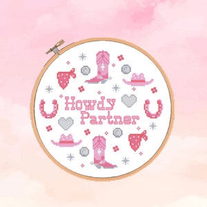 Könnte beinhalten: Ein weißer Stickrahmen mit rosa und grauer Stickerei, die Cowboystiefel, Hüte, Hufeisen, Herzen und Sterne darstellt. Der Text "Howdy Partner" ist in rosa gestickt.