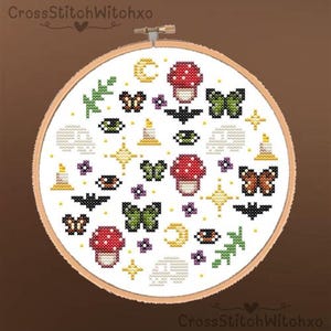 May include: A cross-stitch embroidery hoop with a colourful, whimsical design. The design features mushrooms, butterflies, bats, moons, candles, stars, and floral elements. The text "CrossStitchWitchxo" is visible.