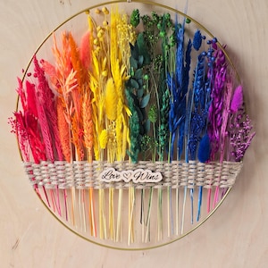 Könnte beinhalten: Ein kreisförmiger Wandbehang aus einem goldenen Metallring mit einem regenbogenfarbenen Arrangement aus gefärbten Trockenblumen und Gräsern. Ein horizontales Band aus geflochtenem Jutegarn fixiert die Stiele, mit einem Holzschild Love Wins.