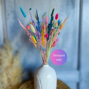 Op de afbeelding: Een levendig boeket van gedroogde grassen en bloemen in tinten turquoise, roze, geel, oranje en blauw, geschikt in een witte keramische vaas met reliëf botanische patronen. Een paarse en roze cirkelvormige afbeelding toont de tekst Bouquet only.