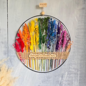 Può includere: Una decorazione da parete circolare con un cerchio in metallo nero riempito da una composizione arcobaleno di fiori ed erbe essiccate. Una fascia intrecciata color cuoio attraversa il centro con una targhetta in legno con scritto "Everyone is welcome here".