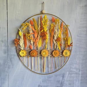 Può includere: Una decorazione da parete circolare con cerchio in metallo dorato che presenta una composizione di fiori ed erbe essiccate nei toni del giallo, arancione e crema. Una fascia intrecciata orizzontale al centro sostiene cinque fiori secchi.