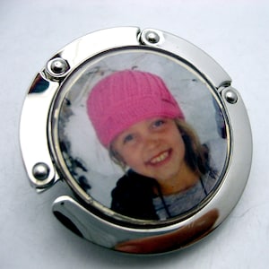 May include: A silver metal purse hook with a round photo insert featuring a young girl wearing a pink knitted hat. The photo is of the girl smiling and looking at the camera.