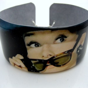 May include: Black and white bangle bracelet with a photo of a woman wearing sunglasses. The woman is looking at the camera with a playful expression.
