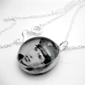 May include: A silver chain necklace with a round pendant featuring a black and white photo of a person wearing a hat.