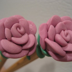 Puede incluir: Dos rosas de tela rosa unidas a palos de madera. Las rosas están hechas de una tela suave y texturizada y tienen un acabado ligeramente brillante.