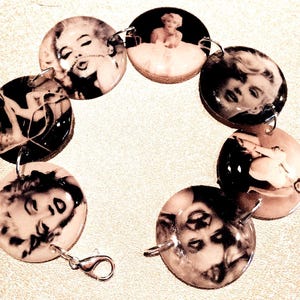 Puede incluir: Un brazalete blanco y negro con dijes redondos que muestran imágenes de Marilyn Monroe. Los dijes están conectados por eslabones de metal plateado.