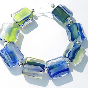 May include: A bracelet composed of rectangular glass beads in shades of blue, lime green, and clear glass, separated by small, round, metallic silver-toned spacer beads strung on a thin, clear, flexible cord arranged in a circular formation.