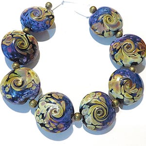 May include: A strand of seven round, flattened glass beads featuring a swirling pattern of deep blue, purple, and cream colors. Each glass bead is separated by a small, metallic brass-toned spacer bead, all strung together on a thin white cord.