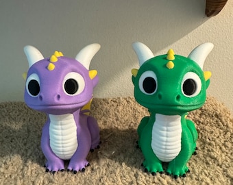 May include: Two 3D-printed dragon figurines. One is purple with yellow accents, the other is green with yellow accents. Both have large black eyes, white horns, and are sitting on a brown, textured surface. These fantasy creatures are decorative items.