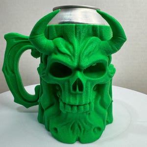 May include: A green, 3D-printed mug with a skull design. The mug features a handle and horns, and the skull's features are clearly defined. The top of the mug holds a silver can. The mug is a unique and decorative item.