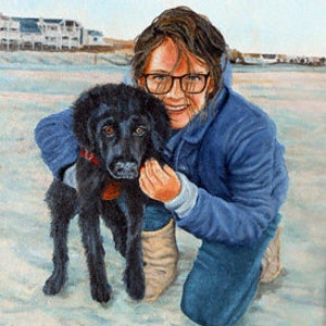 May include: A young person wearing a blue jacket and glasses is kneeling on a sandy beach with a black dog. The dog is looking at the camera and the person is smiling.