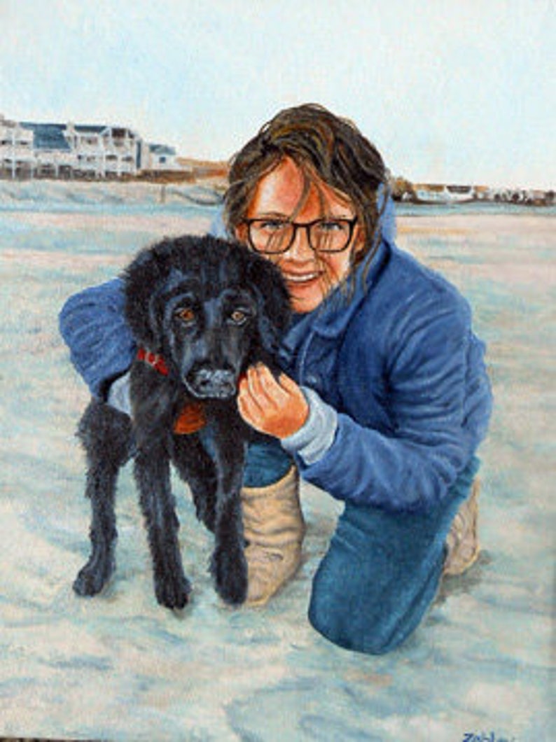 May include: A young person wearing a blue jacket and glasses is kneeling on a sandy beach with a black dog. The dog is looking at the camera and the person is smiling.