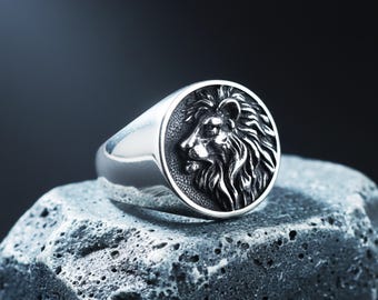 Könnte beinhalten: Ein silberner Siegelring mit einem detaillierten Löwenkopf-Design. Der Ring hat eine runde Oberfläche mit dem Profil des Löwen im Relief. Der Ring wird auf einem grauen, strukturierten Stein vor dunklem Hintergrund präsentiert.