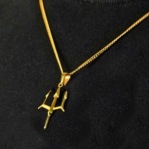 May include: A gold-toned metal necklace featuring a trident-shaped pendant suspended from a delicate curb chain. The pendant has three sharp, pointed prongs and a central vertical shaft, reflecting a bright metallic finish against a dark background.