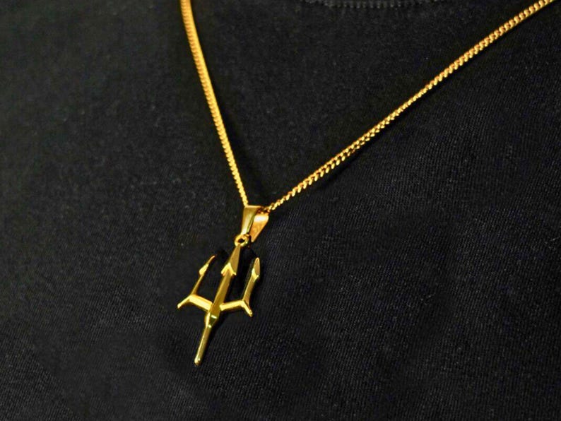May include: A gold-toned metal necklace featuring a trident-shaped pendant suspended from a delicate curb chain. The pendant has three sharp, pointed prongs and a central vertical shaft, reflecting a bright metallic finish against a dark background.