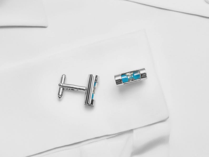 May include: A pair of silver-toned cufflinks featuring a cylindrical design with a clear glass center. Inside the glass, a small amount of bright turquoise sand is visible, creating a miniature hourglass effect within the metal frame.