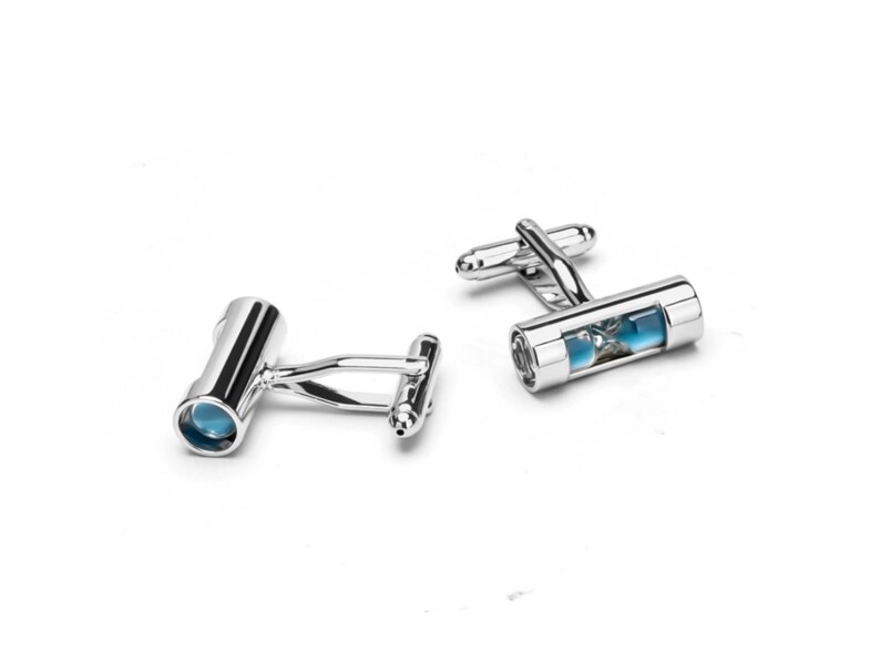 May include: A pair of silver-toned metal cufflinks featuring a cylindrical design with a clear window revealing a small blue hourglass element inside. Each cufflink has a polished finish and a standard hinged toggle closure mechanism on the back.