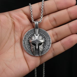 Puede incluir: Un collar con colgante plateado con un dise&ntilde;o detallado de casco espartano. El colgante circular tiene un borde intrincado y cuelga de una cadena a juego. El collar se sostiene en una mano sobre un fondo oscuro.