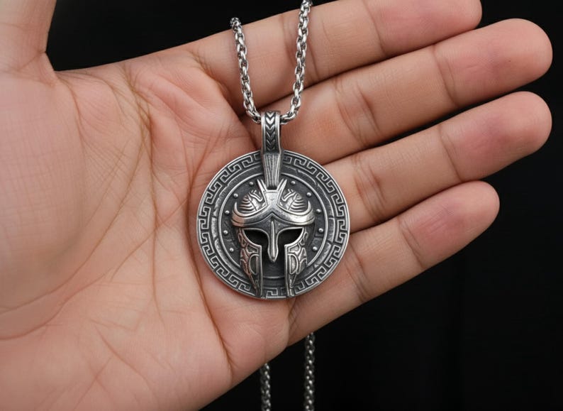Puede incluir: Un collar con colgante plateado con un dise&ntilde;o detallado de casco espartano. El colgante circular tiene un borde intrincado y cuelga de una cadena a juego. El collar se sostiene en una mano sobre un fondo oscuro.