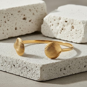 May include: A gold-colored cuff bracelet with seashell-shaped ends. The bracelet is open, with the shell designs facing outward. The bracelet is displayed on a textured, light gray surface, with two white, rectangular blocks in the background.