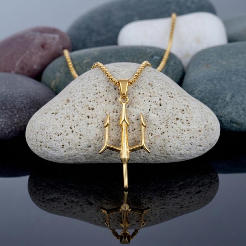 May include: A gold-toned metal trident pendant suspended from a matching gold-toned box chain. The trident features a central vertical prong flanked by two curved side prongs, all connected to a small bail at the top for the chain attachment.