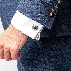 May include: A man wearing a dark blue suit jacket with a white shirt and cufflinks. The cufflinks are round and have a detailed design.