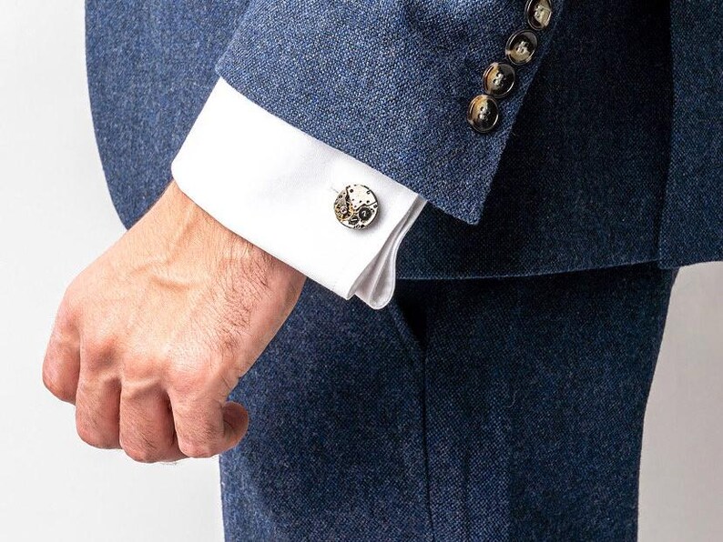 May include: A man wearing a dark blue suit jacket with a white shirt and cufflinks. The cufflinks are round and have a detailed design.