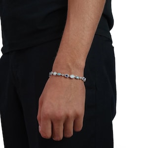 May include: A silver-toned link bracelet featuring alternating oval-shaped white opalescent stones and rectangular blue gemstones. The bracelet is secured with a metal clasp and displays a series of small, clear, sparkling stones set between each larger gem.