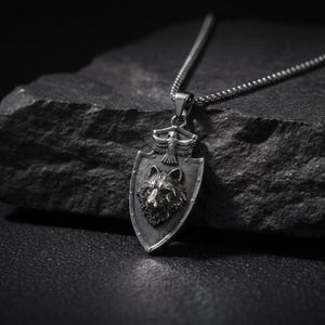 Może przedstawiać: Srebrzysty naszyjnik z wisiorkiem w kształcie tarczy, ozdobiony wypukłym reliefem głowy wilka pośrodku oraz małym emblematem orła u góry. Wisiorek zawieszony jest na cienkim, teksturowanym łańcuszku wykonanym ze srebrzystego metalu.
