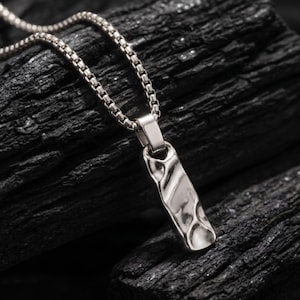 May include: A silver-toned pendant necklace featuring a rectangular charm with an irregular, hammered, and reflective surface texture. The pendant hangs from a thin, flexible box chain, creating a metallic contrast against the dark, rough background.