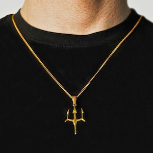 May include: A gold-toned metal necklace featuring a trident-shaped pendant suspended from a thin, delicate chain. The pendant has three distinct prongs and a central vertical shaft, hanging against a plain black fabric background.