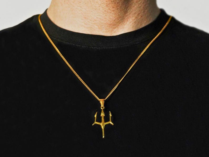 May include: A gold-toned metal necklace featuring a trident-shaped pendant suspended from a thin, delicate chain. The pendant has three distinct prongs and a central vertical shaft, hanging against a plain black fabric background.