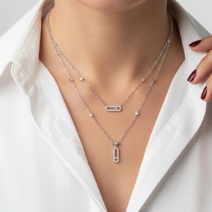 Może przedstawiać: Warstwowy srebrny naszyjnik z błyszczącymi diamentowymi akcentami. Naszyjnik składa się z dwóch sznurów, jeden z poziomym owalnym wisiorkiem, a drugi z pionowym owalnym wisiorkiem. Osoba ma na sobie białą koszulę zapinaną na guziki.