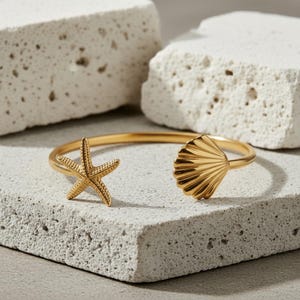 Könnte beinhalten: Ein goldfarbenes, offenes Armband mit einem Seestern- und Muscheldesign. Das Armband wird auf einer strukturierten, hellgrauen Oberfläche mit zwei weißen, rechteckigen Blöcken im Hintergrund präsentiert. Das Armband ist ein Schmuckstück zum Thema Strand.