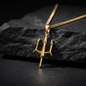 May include: A gold-toned trident pendant necklace featuring a three-pronged design suspended from a matching gold-toned chain. The pendant has a polished finish with sharp, symmetrical points and a central vertical shaft, creating a distinct silhouette.