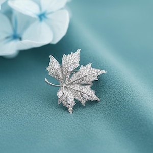 May include: A silver-toned maple leaf brooch, encrusted with sparkling clear crystals. The brooch is set against a teal background, with soft blue flowers in the upper left corner. The brooch is a detailed, nature-inspired accessory.