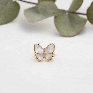 May include: A gold-colored butterfly ring with a white, iridescent center and a line of small, clear stones. The ring's design features a delicate butterfly shape, perfect for adding a touch of nature-inspired elegance to any outfit.
