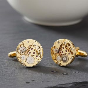 May include: Two gold-coloured cufflinks displaying the inner workings of a watch. The circular cufflinks reveal intricate gears, jewels, and mechanical parts. Set against a dark grey surface, with a white bowl in the background.