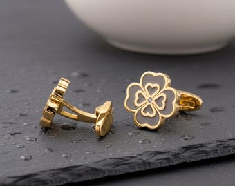 May include: A pair of gold-toned cufflinks featuring a four-leaf clover design. Each clover has a smooth gray inlay with a central heart-shaped cutout pattern and a small, clear embedded stone at the very center of the clover face.