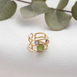 Op de afbeelding: Een goudkleurige ring met een open ontwerp, met drie gekleurde edelstenen: lichtblauw, lichtgroen en roze. De ring staat tegen een witte achtergrond met groene bladeren op de achtergrond.